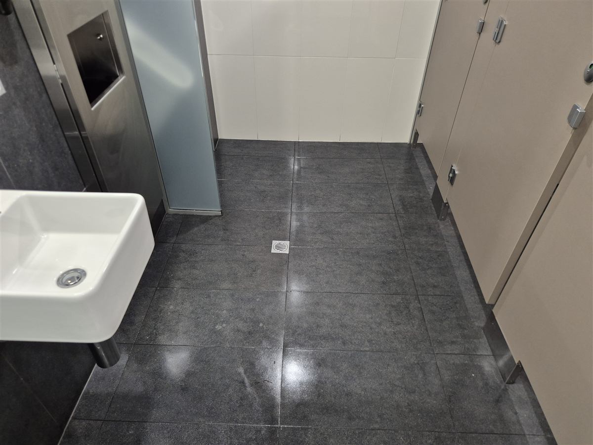 The same washroom after floor cleaning, with the grey tiles and central drain visible between the basin and cubicles