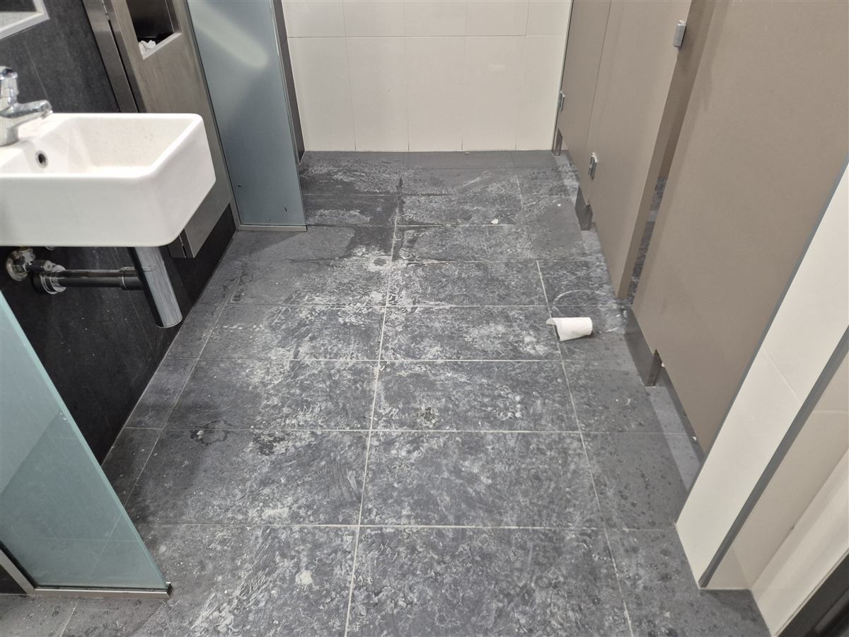 Footprints and pale residue across a washroom floor before cleaning, with a basin on the left and cubicles on the right